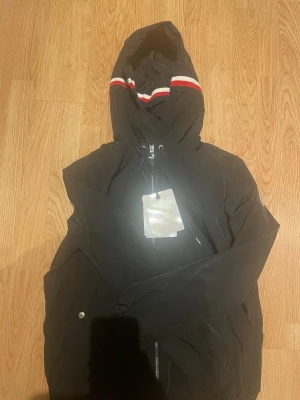 Svart vindjacka från Moncler - Snygg svart vindjacka från Moncler med huva och dragkedja. Jackan har röda och vita ränder på huvan för en sportig look. Tillverkad i lätt och slitstarkt material, perfekt för blåsiga dagar. Klassisk passform och minimalistisk design. 