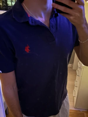 Marinblå pikétröja från US Polo Assasin.  - Säljer en klassisk marinblå pikétröja från US polo assasin. Med röd broderad logga på bröstet. Tröjan har korta ärmar, krage och knappar vid halsen. Perfekt till jeans eller shorts för en clean och stilren look.