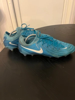 Blå Nike fotbollsskor med snörning - Säljer ett par blå Nike fotbollsskor med vit swoosh och snörning. Skorna har rund tå, platt sula med dobbar och är tillverkade i syntetmaterial med stickad ovandel för extra komfort. Perfekta för dig som vill ha stilrena och funktionella skor på planen. Dem har fortfarande väldigt mycket att ge dem har inga problem. Jag tar gärna emot byte fotbollskor som är i samma storlek