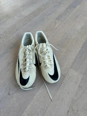 Nike vita fotbollsskor med svart swoosh - Säljer ett par vita Nike fotbollsskor med stor svart swoosh på sidan och coola grafiska detaljer. Skorna har snörning och är tillverkade i syntetmaterial för bra grepp och känsla på planen. Perfekta för dig som vill sticka ut på matchen!