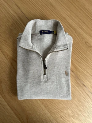 Grå quarter zip från Polo Ralph Lauren - Snygg ljusgrå tröja med quarter zip från Polo Ralph Lauren. Klassisk design med broderad logga på bröstet och hög krage. Tillverkad i mjukt material som känns skönt mot huden. Perfekt för lager-på-lager och enkel att matcha.