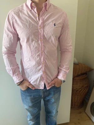 Rosa skjorta från Ralph Lauren - Klassisk rosa skjorta från Ralph Lauren med button-down krage och broderad logga på bröstet. Skjortan har lång ärm, knappar framtill och är tillverkad i bomull. Perfekt för en clean och stilren look. Skick 7/10 storlek M, inga fläckar eller hål