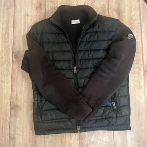 Svart dunväst från Moncler - Snygg svart dunväst från Moncler med stickade mörka ärmar och klassisk Moncler-logga på ena ärmen. Västen har dragkedja framtill och två dragkedjeförsedda fickor. Perfekt för lager-på-lager och streetstyle.