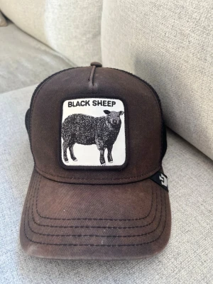 Brun truckerkeps Black Sheep - Säljer en brun truckerkeps från Goorin Bros med ett stort tygmärke framtill där det står 'Black Sheep' och en svart får-illustration. Kepsen har svart mesh baktill och justerbar snapback. 