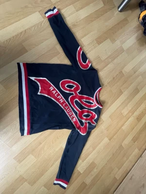 Mörkblå Ralph Lauren tröja - Snygg mörkblå tröja från Ralph Lauren med röda och vita detaljer. Stor logga och text framtill, samt ränder på ärmar och nederkant. Långärmad modell med sportig vibe och rund halsringning.