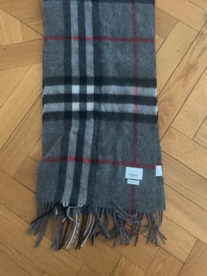 Grå rutig halsduk från Burberry - Snygg grå halsduk från Burberry i klassiskt rutigt mönster med svarta, vita och röda ränder. Tillverkad i 100% kashmir och har fransar i ändarna. Perfekt accessoar för att lyfta din outfit med en lyxig touch.