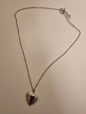 Silverfärgat hjärthalsband med medaljong - Säljer ett stilrent halsband i silverfärg med ett hjärtformat hängsmycke som går att öppna som en medaljong. Kedjan är tunn och smidig, och hjärtat har en blank yta med fina detaljer runt kanten. Perfekt för att lägga i ett litet foto eller minne.