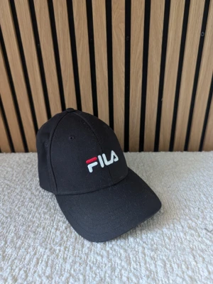 Svart keps från FILA med broderad logga - Snygg svart keps från FILA med böjd skärm och broderad logga i vitt och rött framtill. Klassisk sexpanelsdesign med ventilationshål och justerbar rem bak. Tillverkad i slitstark bomull som passar perfekt till streetwear-looken.