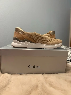 Beige sneakers från Gabor - Stilrena beige sneakers från Gabor med stickad ovandel och elastisk mudd för enkel påtagning. Skorna har detaljer i guld och mocka, samt en vit chunky sula som ger en modern look. Perfekta för dig som gillar bekväma och trendiga skor.