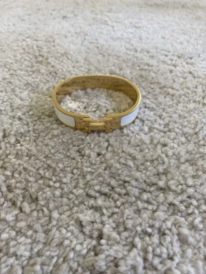 vitt och guldigt armband - Snyggt armband i vitt och guldigt metall. Armbandet har den ikoniska H-detaljen framtill och en stilren, elegant design. Perfekt accessoar för att lyfta din outfit med en lyxig touch. Materialet är metall med emaljinslag.