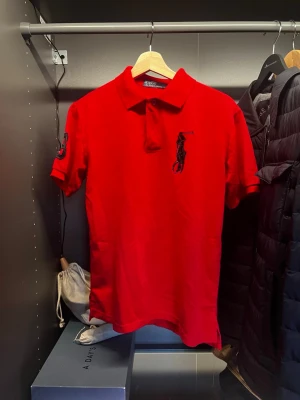 Röd pikétröja från Ralph Lauren - Klassisk röd pikétröja från Ralph Lauren med stor svart logga på bröstet och broderad detalj på ärmen. Tröjan har krage, knappar vid halsen och korta ärmar. Perfekt för en sportig och stilren look.