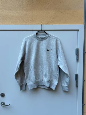 Vintage 1990s nike crewneck - Säljer en stilren grå sweatshirt från Nike med klassisk svart Swoosh-logga på bröstet. Tröjan har rund halsringning, ribbade muddar vid ärmslut och nederkant samt en relaxed fit. Perfekt för dig som gillar sportig och enkel stil.
