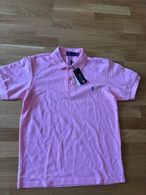 Rosa pikétröja från Polo Ralph Lauren - Snygg rosa pikétröja från Polo Ralph Lauren med klassisk krage och två knappar framtill. Tröjan har det ikoniska blå broderade logotypen på bröstet och är tillverkad i mjuk bomull. Perfekt för en avslappnad men stilren look.