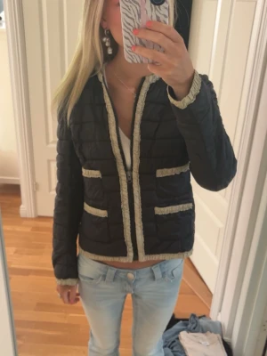 Svart dunjacka med detaljer - Snygg svart pufferjacka med beige fransdetaljer längs kanter, fickor och ärmslut. Jackan har två framfickor och dragkedja framtill. Quiltad design och rund halsringning ger en trendig look. Perfekt för kalla dagar och enkel att styla till olika outfits. Ens fickan är sönder men går lätt att sy tillbaka.  