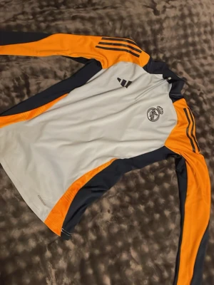 Real Madrid träningströja Adidas - Snygg långärmad träningströja från Adidas med Real Madrid-logga. Tröjan är vit med mörkblå och orange detaljer, har halv dragkedja och klassiska Adidas-ränder på axeln. Perfekt för fotbollsträning eller sportiga aktiviteter.