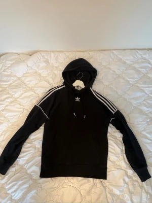 Svart Adidas hoodie med vita ränder - Säljer en svart hoodie från Adidas med klassiska vita ränder längs axlarna och ärmarna. Tröjan har huva med dragsko och Adidas-logga på bröstet. Tillverkad i mjukt material, perfekt för en avslappnad och sportig stil.