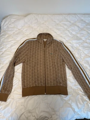 Brun ziptröja från Lacoste - Snygg brun ziptröja från Lacoste med monogrammönster över hela plagget. Tröjan har vita och blå ränder längs ärmarna, ribbade muddar och krage samt det klassiska Lacoste-märket på bröstet. Perfekt för en sportig och trendig look.