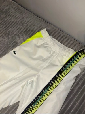 Vita mjukisbyxor från Lacoste - Snygga vita mjukisbyxor från Lacoste med elastisk midja, gula detaljer vid fickorna och en cool svart-grön mönstrad rand längs benet. Klassisk Lacoste-logga på låret. Perfekta för en sportig och avslappnad stil.