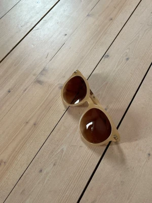 Beige solglasögon med bruna glas - Snygga solglasögon i beige plast med rundad form och bruna tonade glas. Bågarna har små guldfärgade nitar vid hörnen för en extra detalj. Perfekta för soliga dagar och ger en cool retrovibe. Använda fåtal gånger men inga defekter eller tecken på användning. Pris kan diskuteras!🥰