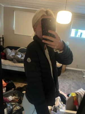 Svart dunjacka från Moncler - Snygg svart dunjacka från Moncler med klassisk quiltad design och hög krage. Jackan har dragkedja framtill och Moncler-logga på ärmen. Perfekt för kalla sommarkvällar eller nått liknade 