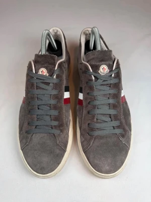 Bruna gråa Moncler sneakers i mocka - Snygga grå bruna sneakers från Moncler i mocka med klassiska Moncler-loggan på plösen och röd, vit och blå rand på sidan. Skorna har grå snörning och vit sula. Perfekta för dig som gillar stilrena och exklusiva snygga sneakers. Bra skick