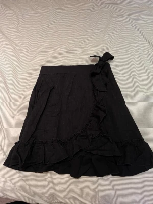 Omlottkjol med volang - Säljer en svart omlottkjol från Gina Tricot i mjukt tyg. Kjolen har volangkant längs nederdelen och knyts i midjan med en rosett. 