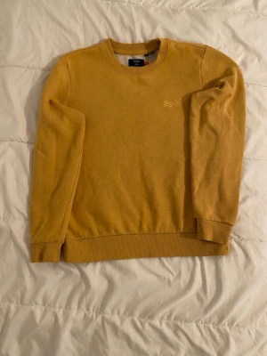 Gul sweatshirt från Superdry - Säljer en gul sweatshirt från Superdry med broderad logga på bröstet. Tröjan har rund halsringning, ribbade muddar vid ärmslut och nederkant samt en avslappnad passform. Perfekt för dig som gillar en enkel men snygg stil.