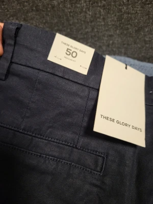Mörkblå chinos från These Glory Days 50 M - Snygga mörkblå chinos från These Glory Days i rak passform. Byxorna har klassiska fickor fram och bak, bälteshällor och stängs med knapp och dragkedja. Perfekta för en clean och stilren look. Materialet känns mjukt och bekvämt.