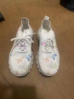 Vita sneakers med fjärilsmönster - Unika vita sneakers med färgglada fjärilar tryckta över hela skon. Skorna har en chunky sula och är tillverkade i ett stickat material som ger en sportig och modern look. Perfekta för dig som vill sticka ut med en lekfull och trendig stil.