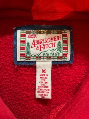 Abercrombie & fitch Vintage - Det står M men passar Xs-S. Säljer nu denna supersnygga och klassiska hoodie från Abercrombie & Fitch i en riktigt härlig röd färg! Tröjan har den ikoniska broderade loggan över bröstet i vitt och marinblått, samt en liten logga på huvan. Den har en perfekt vintage/Y2K-vibe och är gjord i ett rejält, mjukt material som håller värmen bra. Raglanärmarna ger den en snygg passform. • Skick: Mycket fint begagnat skick, väl omhändertagen! • Färg: Klarröd med vita detaljer.