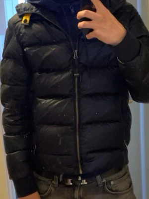 Svart pufferjacka från Parajumpers - Snygg mörkblå pufferjacka från Parajumpers med huva och dragkedja framtill. Jackan har ribbade muddar, två sidofickor med dragkedja och den klassiska gula detaljen vid halsen. Perfekt för kalla vinterdagar och riktigt skön att bära.