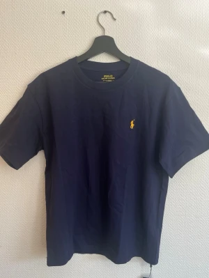 Marinblå t-shirt från Polo Ralph Lauren - Snygg marinblå t-shirt från Polo Ralph Lauren med klassisk gul logga broderad på bröstet. Modellen är slim fit och har rund halsringning samt korta ärmar. Tillverkad i mjuk bomull som känns skön mot huden.