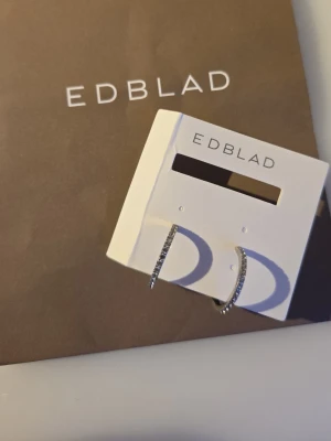 Oanvända edblad örhängen: Glow Creoles Mini M - Snygga silverfärgade örhängen från Edblad i halvbåge med små gnistrande stenar längs framsidan. Tillverkade i rostfritt stål och har en stilren, modern design som passar perfekt till din smyckessamling.