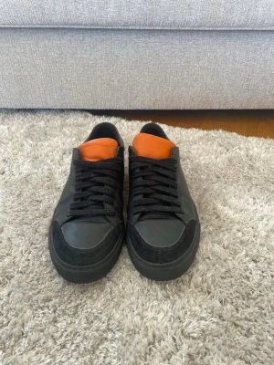 Svarta sneakers från Aregato - Snygga svarta sneakers från Aregato med orange plös och svarta skosnören. Skorna har en låg profil och detaljer i både läder och mocka. På sidan finns texten '1.400MI' i vitt. Perfekta för dig som gillar stilrena och unika sneakers.