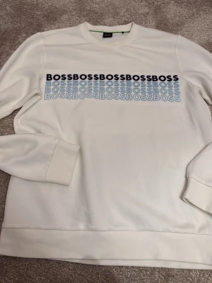 Vit sweatshirt från BOSS - Snygg vit sweatshirt från BOSS med tryckt logga i svart och blått över bröstet. Helt ny inga fläckar eller liknande