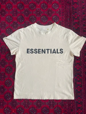 Essentials beige t-shirt - Snygg beige t-shirt från Essentials med svart tryck på bröstet. Klassisk rund halsringning och korta ärmar. Tillverkad i mjuk bomull som känns skön mot huden. Perfekt för en clean och stilren look.