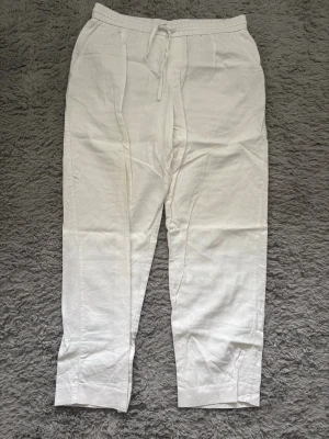 Vita linnebyxor från Zara - Säljer ett par vita linnebyxor från Zara med avslappnad passform och dragsko i midjan. Byxorna har raka ben och resår i midjan för extra komfort. Perfekta för en chill och stilren look under varma dagar.