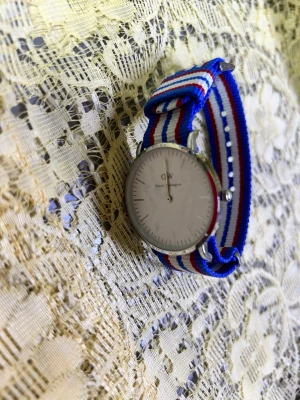 Randig klocka  - Stilren klocka  med vitt urtavla och silverfärgad ram. Armbandet är randigt i blått, vitt och rött och tillverkat i syntetmaterial. Perfekt för dig som vill ha en klassisk och färgglad accessoar.