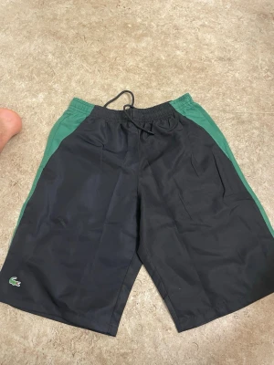 Svarta och gröna shorts från Lacoste - Säljer ett par svarta shorts från Lacoste med gröna sidor och klassisk krokodillogo på vänster ben. Shortsen har elastisk midja med snörning och är tillverkade i ett lätt och sportigt material, perfekta för sommaren eller träning.