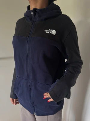 Blå hoodiejacka från The North Face - Mörkblå hoodiejacka från The North Face med svarta axelpartier och vit logotyp på bröst och rygg. Jackan har dragkedja framtill, huva och är tillverkad i mjukt bomullsmaterial. Perfekt för chill dagar och enkel att matcha med jeans eller joggers.