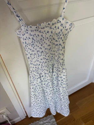 Vit blommig sommarklänning - Söt vit klänning med blått blommönster och smala axelband. Klänningen har smock upptill och faller i en lätt, luftig form. Perfekt för varma dagar och har en romantisk vibe. Materialet känns som bomull eller linne.