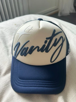Vanity truckerkeps blå/vit - Truckerkeps, krämvit och navy blå färg. Puff print på framsidan med broderat text på sidan