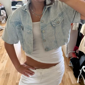 Cropped ljusblå jeansjacka/bolero med rosetter - Säljer en cropped ljusblå jeansjacka/bolero med korta ärmar och coola rosettdetaljer i ryggen. Jackan har två bröstfickor med knappar och en avslappnad passform. Perfekt för dig som vill sticka ut med en unik jeansjacka. Den har en liten fläck som man ser på bild ett men inget man tänker på!