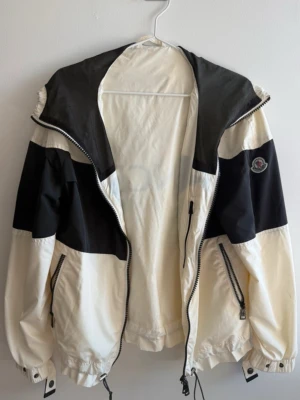 Moncler svartvit vindjacka med huva - Snygg vindjacka från Moncler i svart och vitt med huva, dragkedja och två fickor med zip. Jackan har meshpanel på ryggen och justerbara snören vid nederkant och ärmslut. Moncler-logga på ärmen. Perfekt för en cool och sportig look.