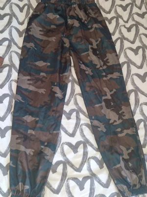Camo mjukisbyxor med resår - Säljer ett par mjukisbyxor med camouflagemönster i grönt, brunt och svart.  Inte riktigt mjukisbyxor material-men fortfarande sköna, och tunna. Byxorna har resår i midjan och vid bensluten för en skön och avslappnad passform. Perfekta för chill dagar eller när du vill ha en streetwear-look. Använd en gång