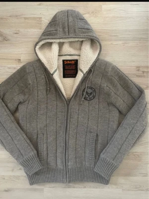 Grå cardigan från schott storlek s - Mysig grå hoodie från Schott med dragkedja, ribbade muddar och två fickor framtill. Insidan är fodrad med vit teddy för extra värme. Märke på bröstet och justerbar huva med snören. Perfekt för kalla dagar och riktigt snygg streetstil.