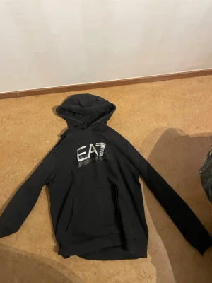 Svart EA7 hoodie från Emporio Armani - Snygg svart hoodie från Emporio Armani med stort EA7-tryck i vitt på bröstet. Klassisk huva och känguruficka framtill. Perfekt för dig som gillar streetwear och vill ha en stilren look med exklusiv känsla., står xxl men passar till L-M