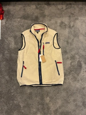 väst från Patagonia - Säljer en beige fleeceväst från Patagonia med marinblå detaljer och dragkedjor. Västen har två fickor med röda dragkedjor och en hög krage. Materialet är mjukt och fluffigt, perfekt för lager-på-lager. Klassisk Patagonia-logga på bröstet.