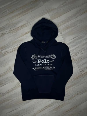 Mörkblå hoodie från Polo Ralph Lauren - Snygg mörkblå hoodie från Polo Ralph Lauren med stor vit logga och text på bröstet. Klassisk huva med dragsko och magficka framtill. Tillverkad i mjukt bomullsmaterial som känns skönt mot huden. Perfekt för en avslappnad och trendig look.