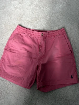 Rosa shorts från Polo Ralph Lauren - Säljer ett par klassiska rosa shorts från Polo Ralph Lauren med elastisk midja, knapp och dragkedja. Shortsen har sidofickor och en liten broderad logga i mörkblått på benet. Perfekta för sommaren och riktigt sköna att bära.